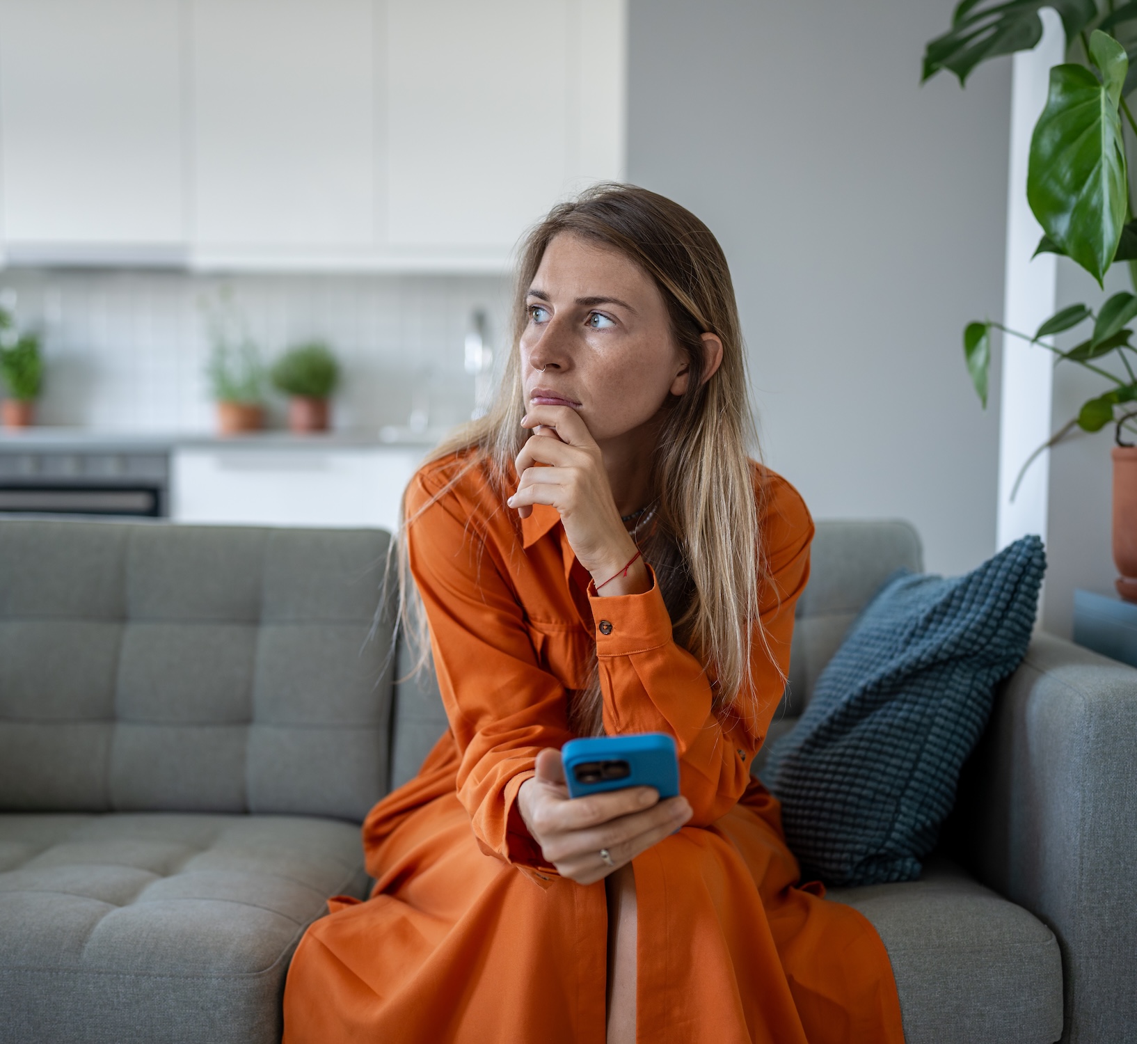 Thoughtful serious woman waits for message or call on phone, sitting alone on couch at home